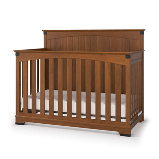 The Child Craft Redmond Full Panel 4-in-1 Convertible Crib in Coach Cherry features vertical slats, a high headboard, black corner accents, and includes a white mattress—ideal for a stylish nursery.