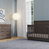 A modern nursery with blue walls, a dark wooden crib, a matching Child Craft Redmond 3 Drawer Dresser in Dapper Gray, a floor lamp with a round shade, white-curtained window, blue rug, and abstract wall art.