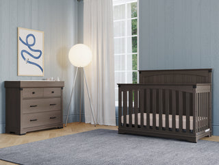 A modern nursery with blue walls, a dark wooden crib, a matching Child Craft Redmond 3 Drawer Dresser in Dapper Gray, a floor lamp with a round shade, white-curtained window, blue rug, and abstract wall art.