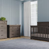 A nursery with light blue walls, a window with white curtains, a gray crib, the Child Craft Redmond 4 Drawer Chest in Dapper Gray as a dresser, a changing table, a gray rug on light wood floors, and a small plant on the chest.