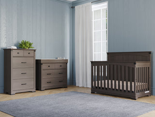 A nursery with light blue walls, a window with white curtains, a gray crib, the Child Craft Redmond 4 Drawer Chest in Dapper Gray as a dresser, a changing table, a gray rug on light wood floors, and a small plant on the chest.
