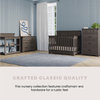 A nursery with dark wood furniture, light blue walls, and a large window with white curtains. Features include a crib, Child Craft Redmond 3 Drawer Dresser in Dapper Gray, changing table, bookshelf, and gray rug on wooden floors. Crafted Classic Quality.
