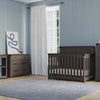 A nursery with light blue walls, hardwood floors, white-curtained window, and dark wood furniture including a crib, dresser, changing table, and a Child Craft Redmond 4 Drawer Chest in Dapper Gray, plus a gray rug.