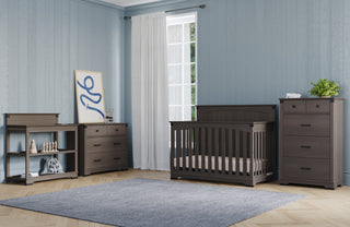 A nursery with light blue walls, hardwood floors, white-curtained window, and dark wood furniture including a crib, dresser, changing table, and a Child Craft Redmond 4 Drawer Chest in Dapper Gray, plus a gray rug.