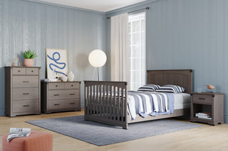 A modern bedroom featuring the Child Craft Redmond Full Panel 4-in-1 Convertible Crib in Dapper Gray, paired with gray wooden furniture, a striped bed set, nightstand, dresser, chest, floor lamp, patterned rug, and sheer curtains.