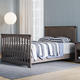 A Child Craft Redmond Full Panel 4-in-1 Convertible Crib in Dapper Gray stands in a cozy bedroom with light blue walls, a tall globe floor lamp, a large window with sheer curtains, and a gray rug on a wooden floor.