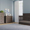 A modern nursery with Child Craft’s Redmond 4 Drawer Chest in Dapper Gray, matching gray crib and dresser, abstract art and plants on the dresser, light blue walls, and a large window with white curtains.