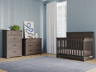A modern nursery with Child Craft’s Redmond 4 Drawer Chest in Dapper Gray, matching gray crib and dresser, abstract art and plants on the dresser, light blue walls, and a large window with white curtains.
