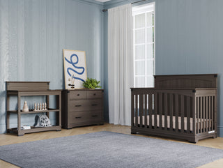 A modern nursery features the Child Craft Redmond Full Panel 4-in-1 Convertible Crib in Dapper Gray, a matching dresser and changing table, light blue walls, large window with sheer curtains, gray rug on wood floor, framed print, and small toys.