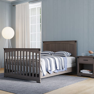 A bedroom featuring the Child Craft Redmond Full Panel 4-in-1 Convertible Crib in Dapper Gray, striped bedding, a matching nightstand, round floor lamp, white-curtained window, gray rug on light wood floor, and light blue walls.