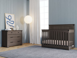 A modern nursery featuring the Child Craft Redmond Full Panel 4-in-1 Convertible Crib in Dapper Gray, a matching dresser, round floor lamp, abstract wall art, decor items, blue paneled walls, light curtains, and a gray rug on wood floors.