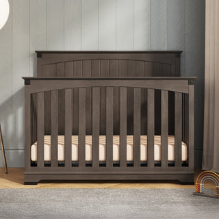 The Child Craft Redmond Full Panel 4-in-1 Convertible Crib in Dapper Gray stands on a light wood floor with a light mattress inside. Against a gray wall, a small rainbow toy is placed to the right of the crib’s curved headboard and vertical slats.