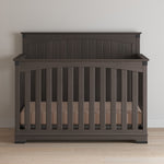 The Child Craft Redmond Full Panel 4-in-1 Convertible Crib in Dapper Gray features vertical slats and an arched headboard, placed against a light wall on a wood floor, with a beige mattress inside.