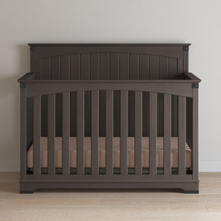 The Child Craft Redmond Full Panel 4-in-1 Convertible Crib in Dapper Gray features vertical slats and an arched headboard, placed against a light wall on a wood floor, with a beige mattress inside.