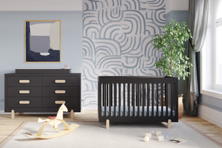 Modern nursery with a black crib and Child Craft Legacy Changing Table Topper in Caviar Black, matching dresser, light wood rocking horse, abstract wall art, potted plant, geometric mural, and sunlight streaming onto a cozy rug.