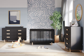 Modern nursery with black furniture, featuring the Child Craft Legacy Changing Table Topper in Caviar Black on a dresser, plus a matching crib and chest of drawers. A wooden rocking horse rests on a light rug, accented by plants, abstract art, and sunlight for coziness.