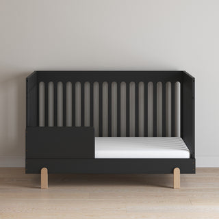 The Child Craft Park Heights 4-In-1 Euro Crib in Caviar Black stands on wooden legs against a light wall, with open side and back slats and a white mattress, featuring a low partial side rail and sleek modern design.
