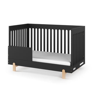 The Child Craft F09505 Toddler Guard Rail in Caviar Black features slatted sides, a guard rail, light wood legs, and a white mattress, all set against a plain white background.