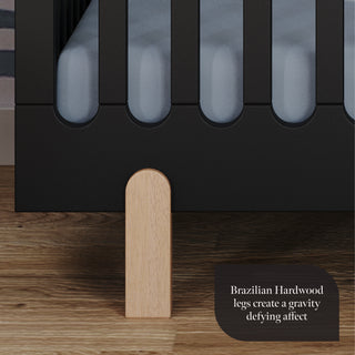Close-up of the Child Craft Park Heights 4-In-1 Euro Crib - Caviar Black, featuring Brazilian hardwood legs on a wooden floor. Text reads: Gravity-defying Brazilian Hardwood legs—ideal for fans of mid-century nursery design.