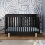 The Child Craft Park Heights 4-In-1 Euro Crib in Caviar Black stands on a wooden floor with a blue abstract wavy pattern on the white wall behind, illuminated by soft light with mid-century nursery accents and playful toys in the foreground.