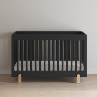The Park Heights 4-In-1 Euro Crib by Child Craft in Caviar Black features wooden legs and slatted sides. Set on a wooden floor against a light wall, this modern crib with a white mattress adds artisan mid-century style to any nursery.