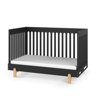 The Child Craft Park Heights 4-In-1 Euro Crib in Caviar Black features open slatted sides and back, a white mattress, and short wooden legs—ideal for a stylish, mid-century nursery set against a white background.