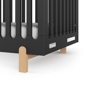 Close-up of the Child Craft Park Heights 4-In-1 Euro Crib in Caviar Black, featuring vertical slats, light wood legs, a white mattress, and logo on the lower panel—perfect for a stylish mid-century nursery.