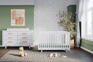 A bright nursery with green and white walls features a white crib, Child Craft Legacy Changing Table Topper in Matte White on a double dresser, a teddy bear, soft blocks on a striped rug, leafy plant by the window, and modern art above the dresser.