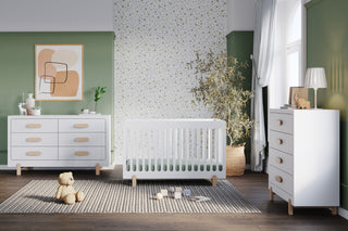 A stylish nursery features a white crib, a Child Craft Park Heights 4-Drawer Chest in matte white with Brazilian hardwood accents, a teddy bear on a striped rug, abstract art, and a potted plant against patterned green walls with sunlight streaming in.