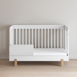 The Child Craft F09505 Toddler Guard Rail in matte white features vertical slats on three sides and wooden legs, shown against a beige wall on a light wood floor.