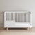 The Child Craft F09505 Toddler Guard Rail in matte white features vertical slats on three sides and wooden legs, shown against a beige wall on a light wood floor.
