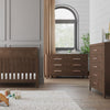 A modern nursery with a wooden crib, Child Craft Kieran 6-Drawer Double Dresser in Toasted Chestnut, chest of drawers, neutral walls, a large window with curtains, indoor plants, and a plush fox toy on a light textured rug.