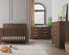 A modern nursery features the Child Craft Kieran 5-Drawer Chest in Toasted Chestnut, paired with a matching wooden crib and dresser. Beige curtains, indoor plants, toys, and a soft rug add cozy touches to the stylish room.