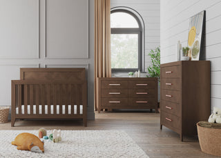 A modern nursery features the Child Craft Kieran 5-Drawer Chest in Toasted Chestnut, paired with a matching wooden crib and dresser. Beige curtains, indoor plants, toys, and a soft rug add cozy touches to the stylish room.