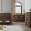 A modern nursery features a wooden crib, the Child Craft Kieran 6-Drawer Double Dresser in Toasted Chestnut, a chest of drawers, beige curtains at the window, a potted plant, and toys on a rug for a cozy touch.