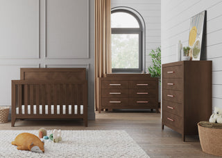Modern nursery room with the Child Craft Kieran 4-in-1 Convertible Crib in Toasted Chestnut, dresser, and chest of drawers. A window with beige curtains, potted plant, toys, and soft rug complete this cozy, bright space.