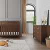 Modern nursery with a wooden crib, Child Craft Kieran 6-Drawer Double Dresser in Toasted Chestnut, plush toys on a rug, a large window with beige curtains, framed picture, plant, teddy bear decor, and light gray walls with white shiplap.