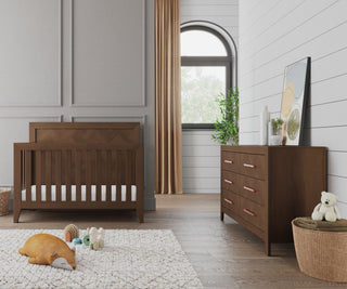 A modern nursery featuring the Child Craft Kieran 4-in-1 Convertible Crib in Toasted Chestnut, a matching dresser with plants and a framed print, plush toys on a textured rug, and natural light through beige curtains.
