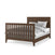 The Child Craft Kieran 4-in-1 Convertible Crib in Toasted Chestnut features vertical slats on the headboard and footboard, and is styled with white and gray bedding and pillows.