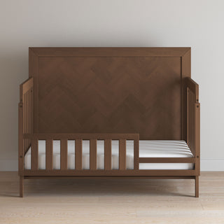 The Child Craft Kieran 4-in-1 Convertible Crib in Toasted Chestnut features a high herringbone-pattern headboard, open side slats, and a white mattress, set against a light wall on a wooden floor.