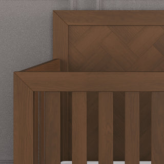 Close-up of the Child Craft Kieran 4-in-1 Convertible Crib in Toasted Chestnut, featuring vertical slats and a herringbone headboard, set against a textured gray wall.