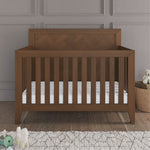 The Child Craft Kieran 4-in-1 Convertible Crib in Toasted Chestnut with a white mattress is on a wooden floor by a textured gray wall, surrounded by toys and a teddy bear in a woven basket.