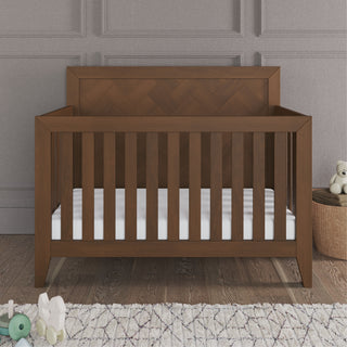 The Child Craft Kieran 4-in-1 Convertible Crib in Toasted Chestnut with a white mattress is on a wooden floor by a textured gray wall, surrounded by toys and a teddy bear in a woven basket.