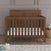 The Child Craft Kieran 4-in-1 Convertible Crib in Toasted Chestnut with a white mattress is on a wooden floor by a textured gray wall, surrounded by toys and a teddy bear in a woven basket.