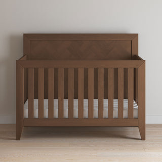 The Child Craft Kieran 4-in-1 Convertible Crib in Toasted Chestnut, featuring vertical slats and a high backboard, stands on a light wooden floor against a plain beige wall. The crib includes a mattress with a white fitted sheet.