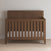 The Child Craft Kieran 4-in-1 Convertible Crib in Toasted Chestnut, featuring vertical slats and a high backboard, stands on a light wooden floor against a plain beige wall. The crib includes a mattress with a white fitted sheet.