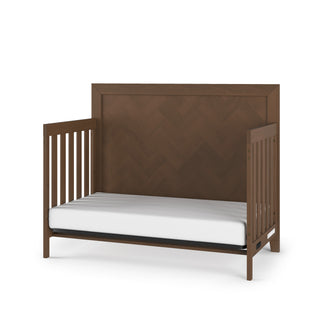 The Child Craft Kieran 4-in-1 Convertible Crib in Toasted Chestnut features a modern herringbone-patterned headboard, open slatted sides, and a white mattress with a minimalistic dark brown finish.