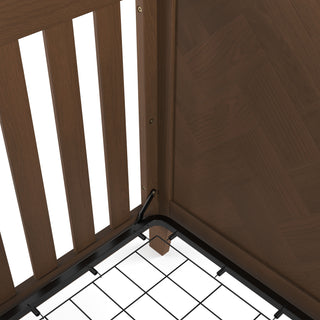 Close-up of the Child Craft Kieran 4-in-1 Convertible Crib in Toasted Chestnut, highlighting its wooden corner with herringbone paneling, side slats, and a black metal grid mattress support frame.