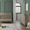 A modern nursery with green walls, wooden crib, and matching dresser topped with the Child Craft F01792 Universal Dressing Kit in Crescent Grey. A round rug with toys covers the floor, and an arched window fills the space with light.