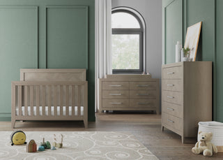 A modern nursery features the Child Craft Kieran 4-in-1 Convertible Crib in Crescent Gray, a dresser, and changing table against green paneled walls. Natural light fills the room, with toys and a teddy bear atop a patterned rug on wood floors.